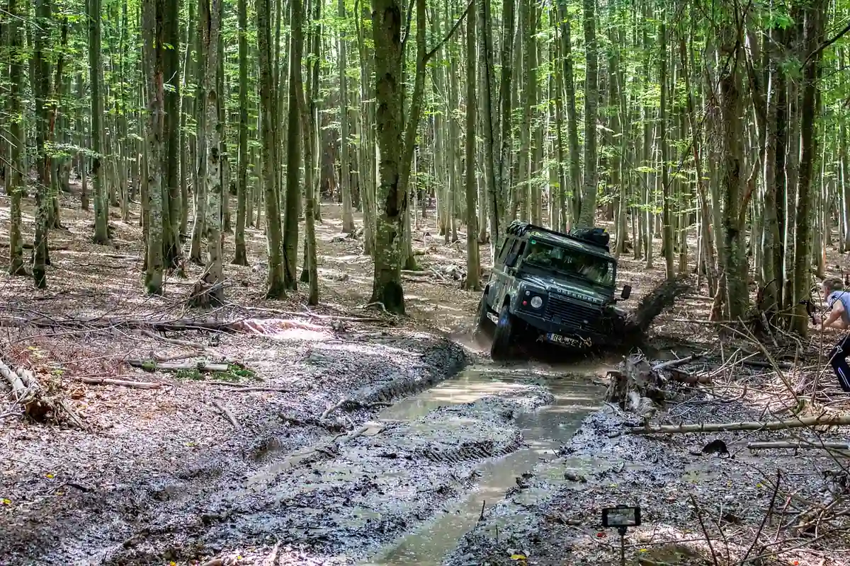A green four-wheel-drive vehicle working through a deep muddy rut on a forest trail while a spotter stands nearby