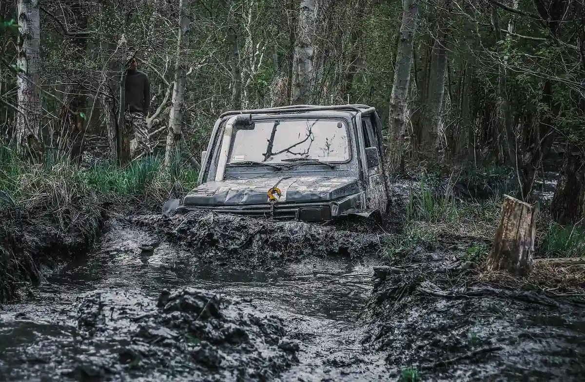 Abandoned-looking 4x4 stuck up to its bumper in a deep mud hole on a forest trail, with trees on both sides