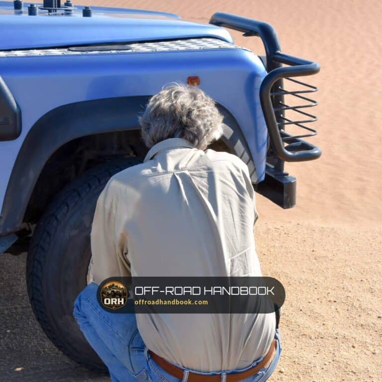 Man kneeling at the front tire of a blue 4x4 in desert sand, adjusting tire pressure — Off-Road Handbook