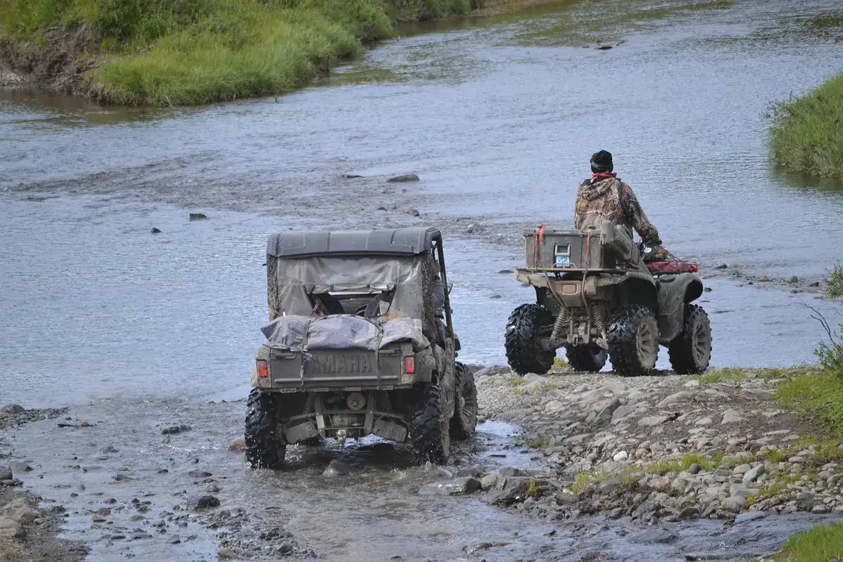 Rider on a utility ATV and a side-by-side crossing a shallow, rocky river