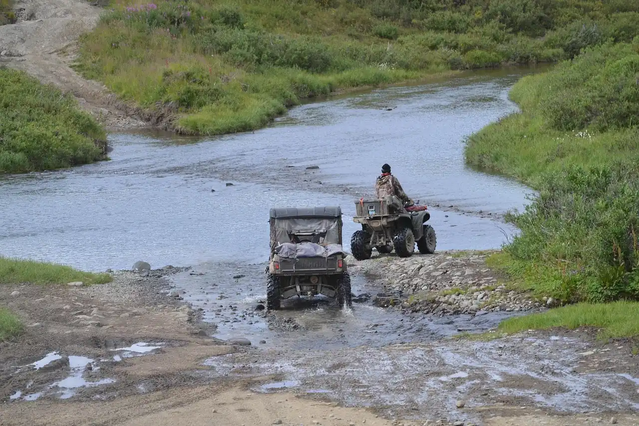 An ATV and a utility vehicle crossing a shallow, muddy stream on a dirt trail