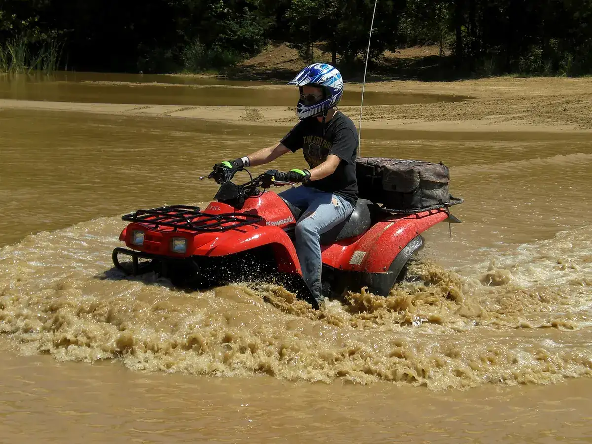 Rider in a helmet driving a red ATV through shallow muddy water with spray on both sides