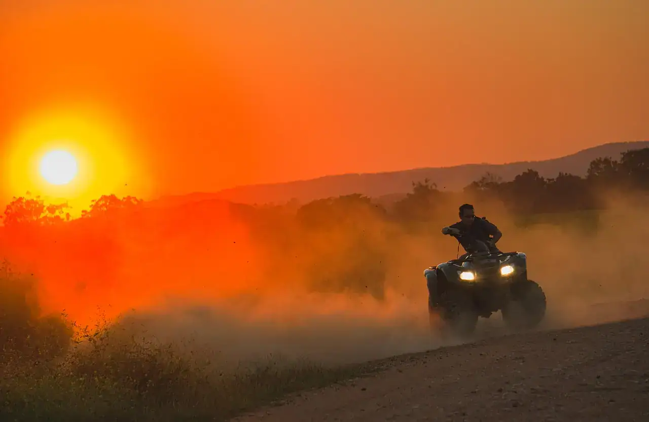 Rider on a four-wheel ATV with headlights on kicking up a thick cloud of dust at sunset