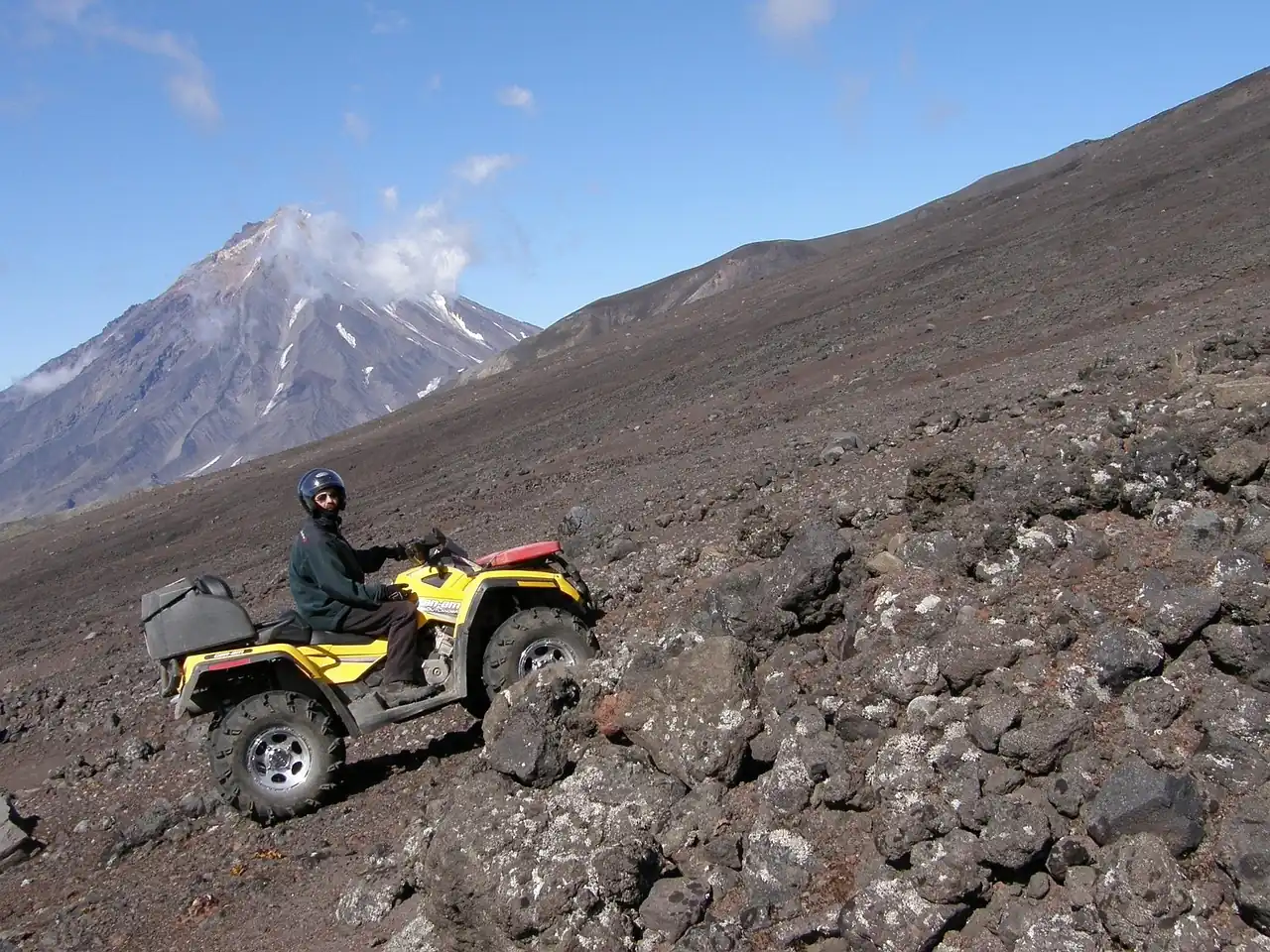 Rider on a yellow ATV climbing a steep slope of dark volcanic rock with a snow-streaked peak behind