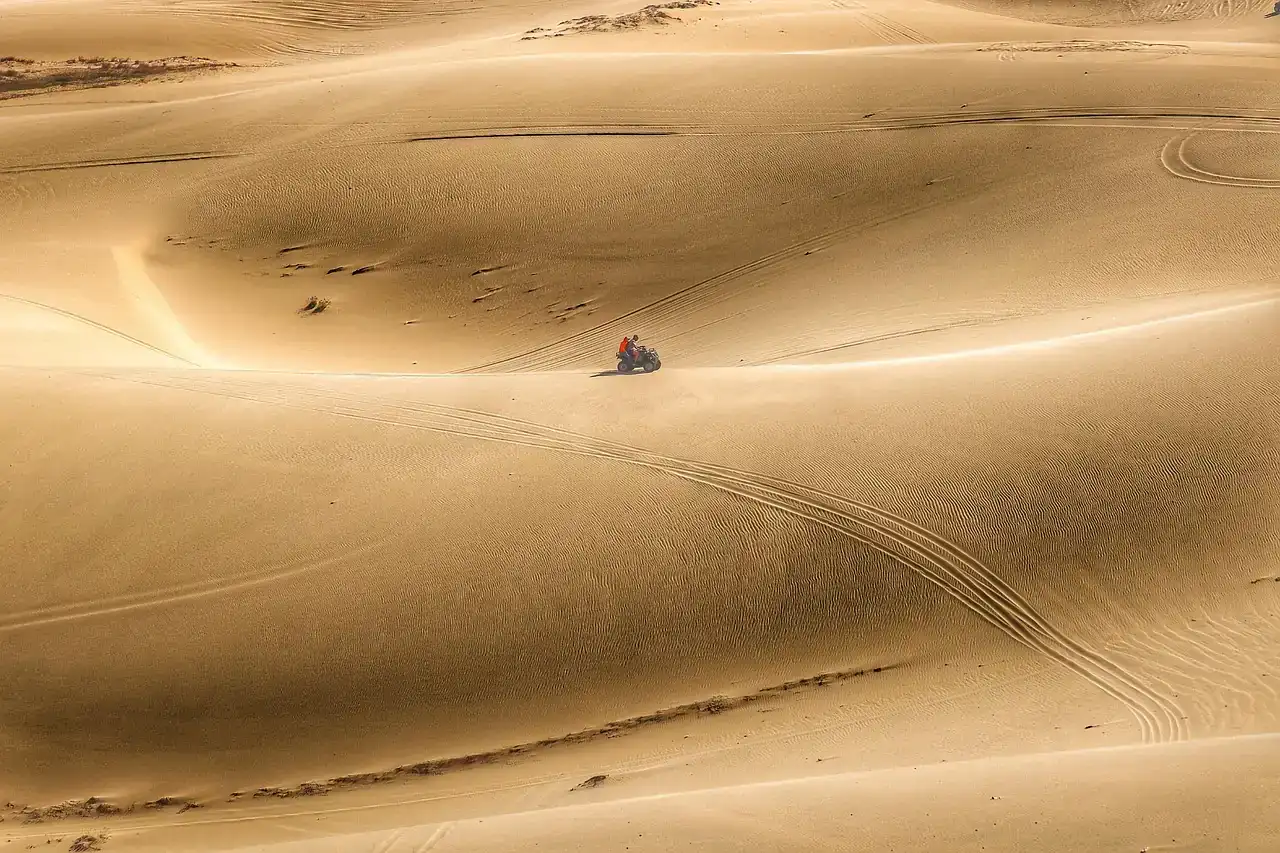 Single ATV rider crossing large sand dunes with tire tracks in the sand