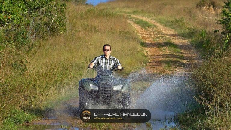 Rider on a utility ATV splashing through a puddle on a grassy trail — Off-Road Handbook