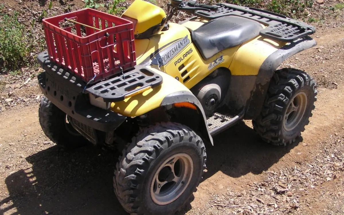 Straddle-seat ATV with front and rear racks, a crate strapped on the rack and knobby tires on a dirt trail