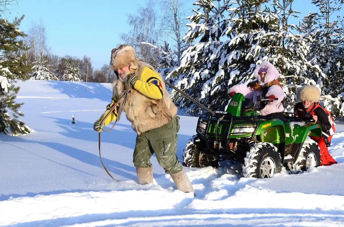 Man pulling a green utility ATV stuck in deep snow with a rope while the riders stay on board