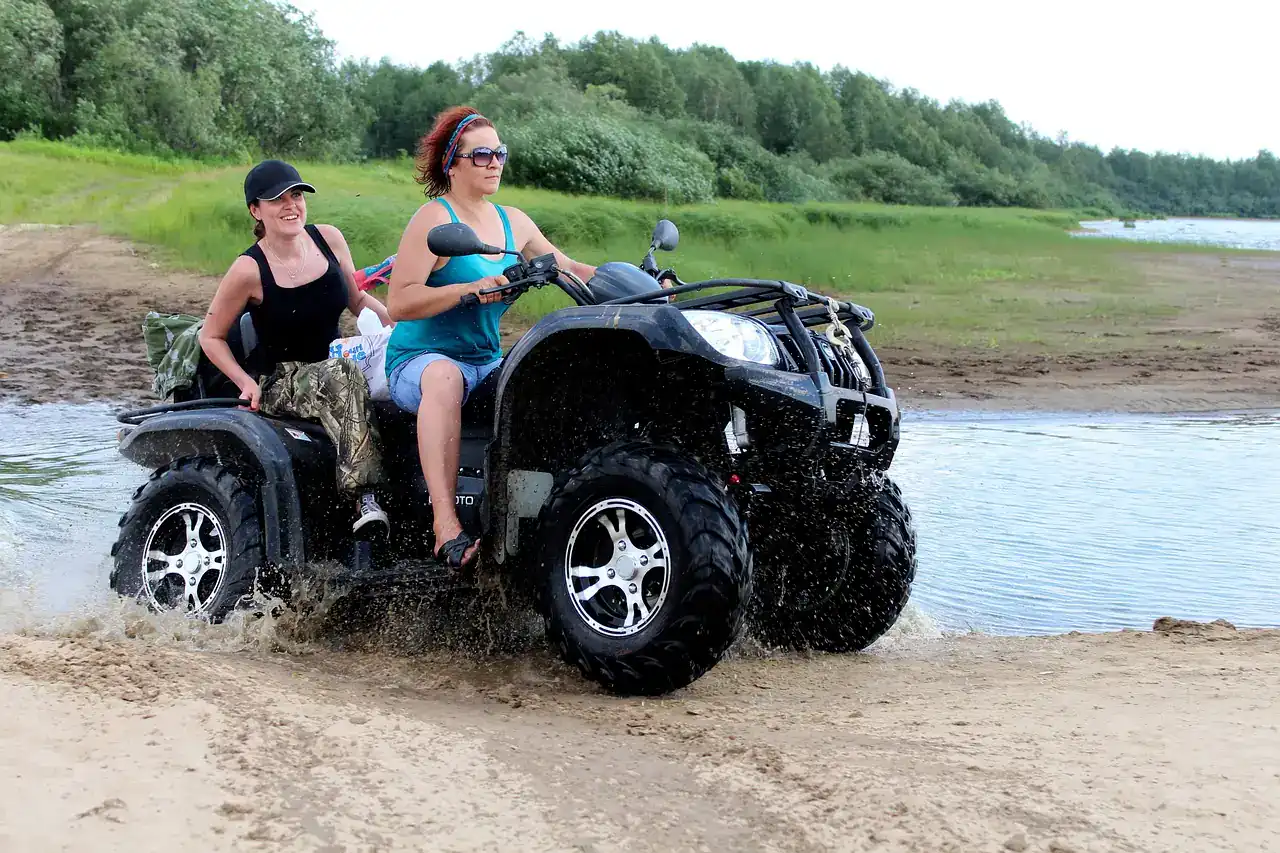 Two riders on a black utility ATV crossing shallow water with spray at the wheels
