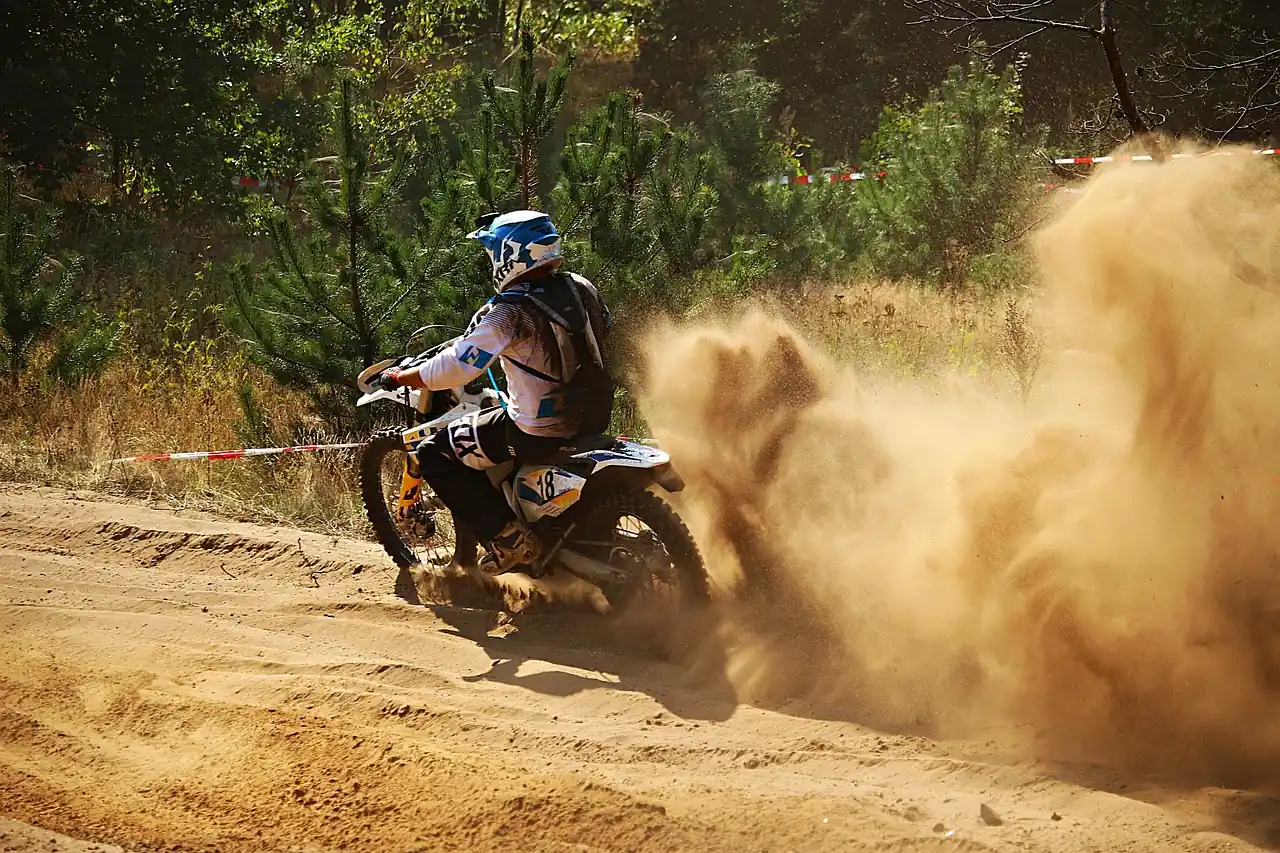 Dirt bike rider kicking up a cloud of dust on a deep sand trail between pine trees