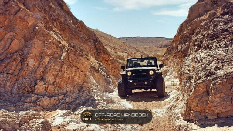Open-top 4x4 with its headlights on driving down a narrow dirt trail between red rock canyon walls