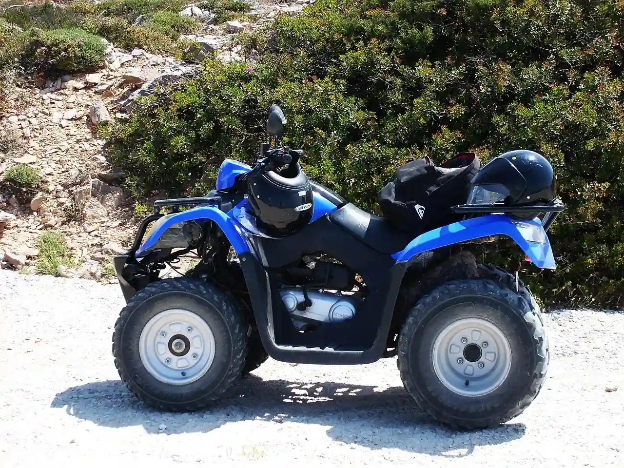 Blue four-wheel utility ATV with a rear case parked on a gravel track beside shrubs