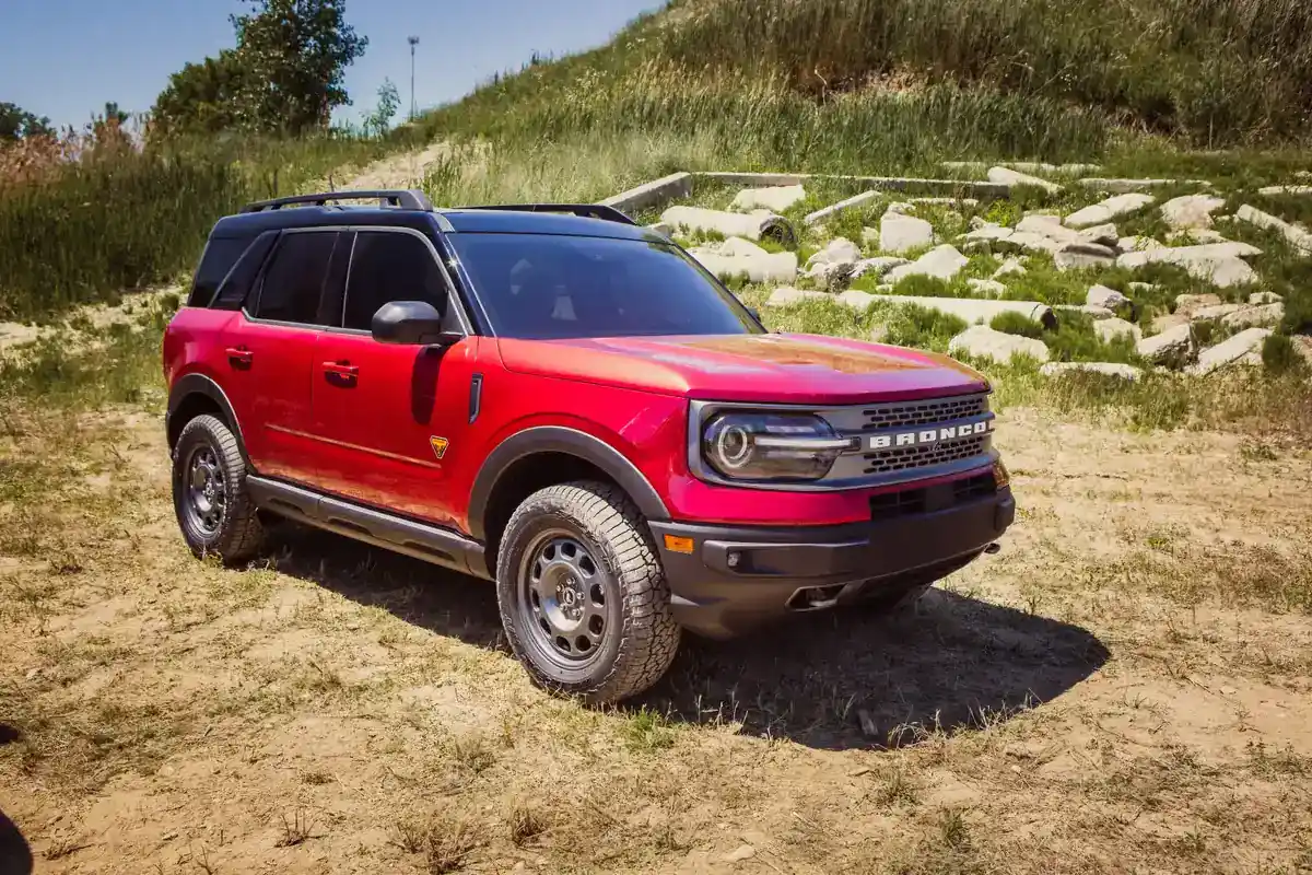 Red Ford Bronco Sport with all-terrain tires and a steel skid plate parked on a dirt and grass hillside beside a rock-lined slope