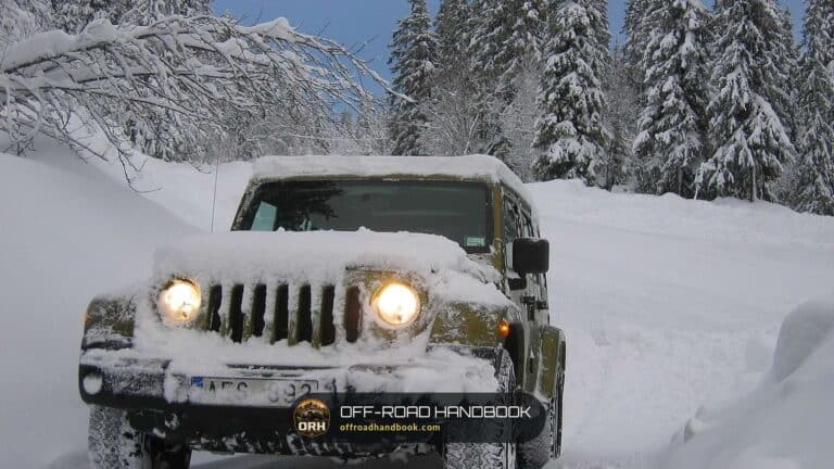 Four-wheel-drive SUV with snow packed on its hood and grille on a snowy forest road, headlights on