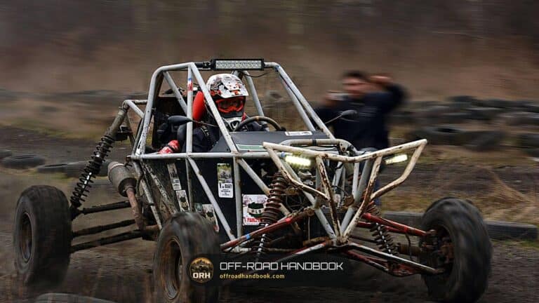 Tube-frame off-road buggy with a driver in a helmet kicking up dirt on a muddy track