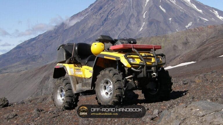 Yellow four-wheel ATV with a front rack and winch parked on a rocky volcanic slope below a snow-streaked peak