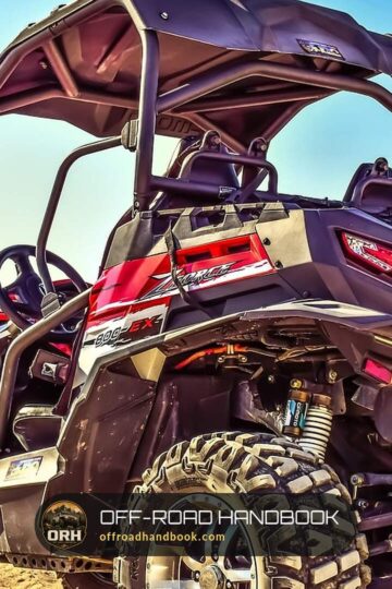 Rear three-quarter view of a red and black side-by-side with roll cage and large all-terrain tires on a sandy trail