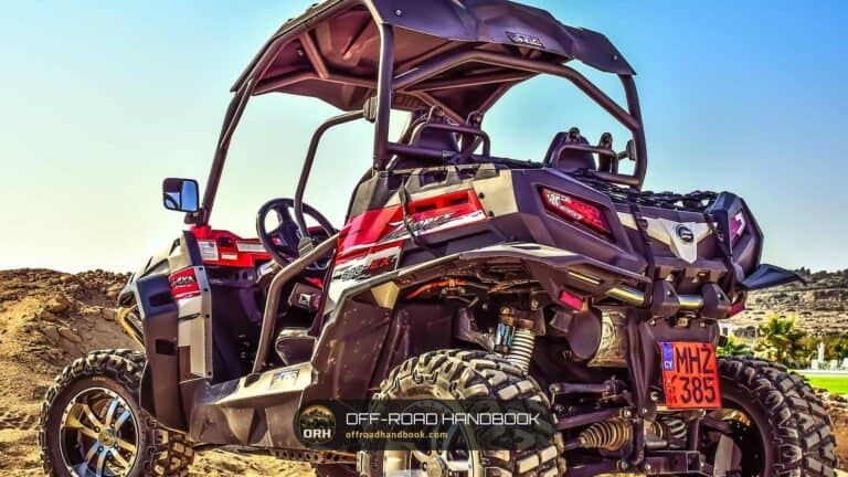 Rear three-quarter view of a red and black side-by-side with roll cage and large all-terrain tires on a sandy trail