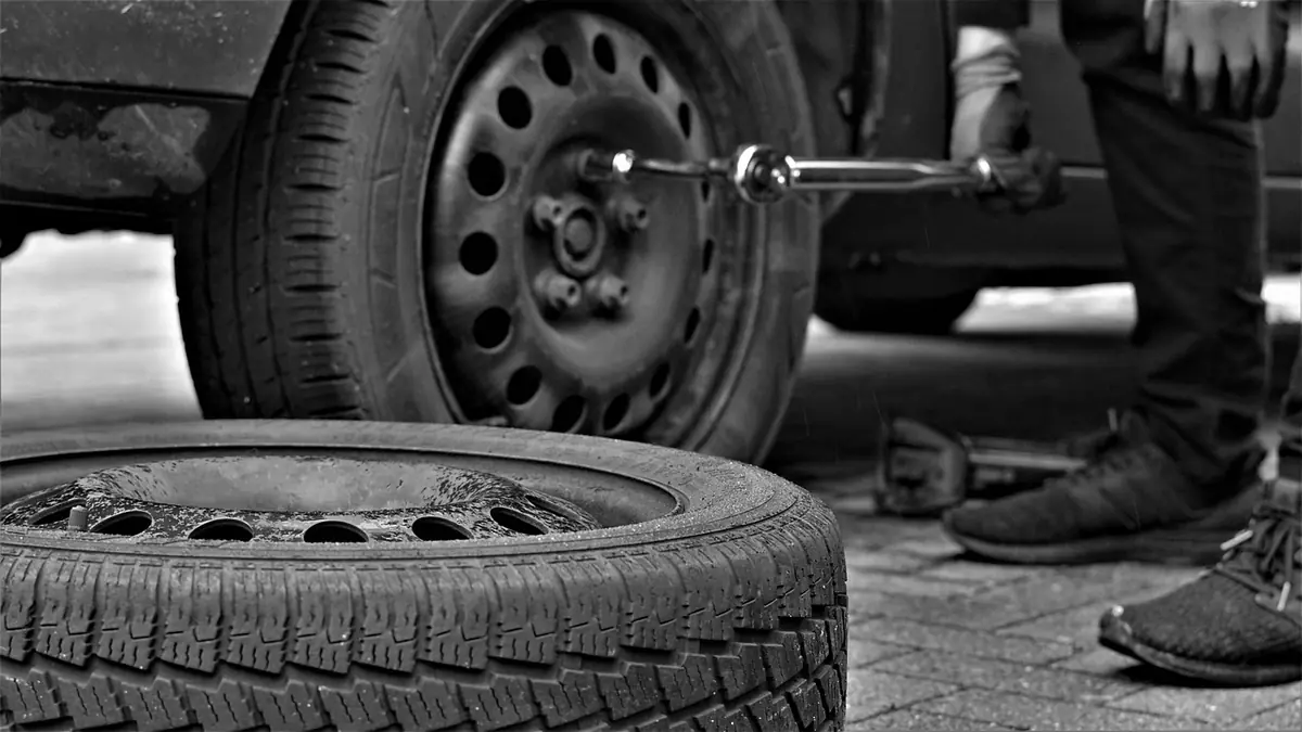 Black and white photo of a person loosening lug nuts on a steel wheel with a lug wrench, spare tire on the ground