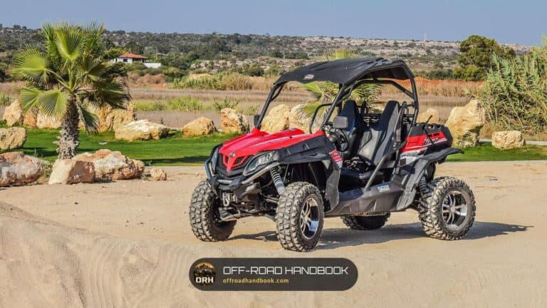 Red and black side-by-side UTV with roll cage parked on sand with rocks and a palm tree behind it