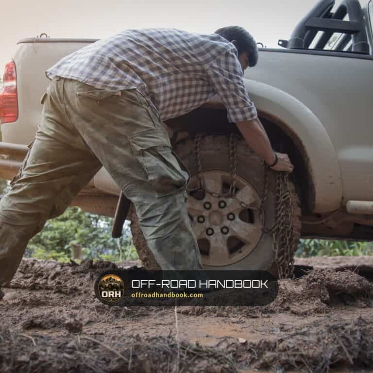 Driver bent over the muddy rear wheel of a silver pickup truck on a dirt trail — Off-Road Handbook