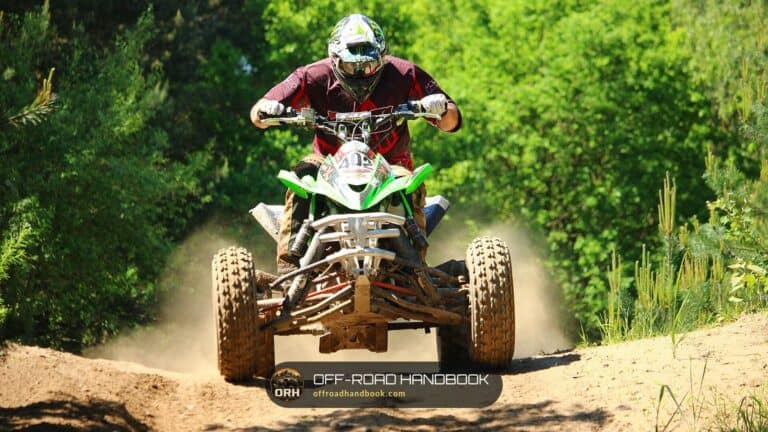 Rider on a green sport ATV kicking up dust on a dirt trail with trees behind