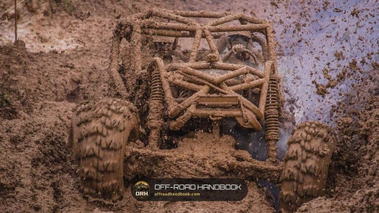 Mud-covered open-frame buggy plowing through deep mud with mud flying