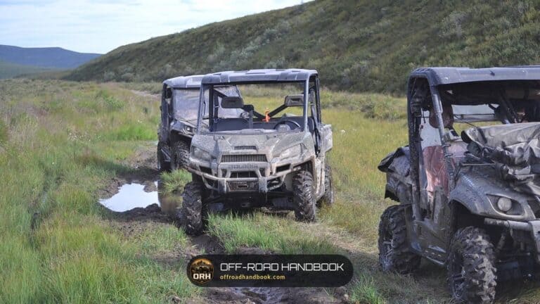 Two muddy side-by-side UTVs stopped on a wet grassy trail below a hillside