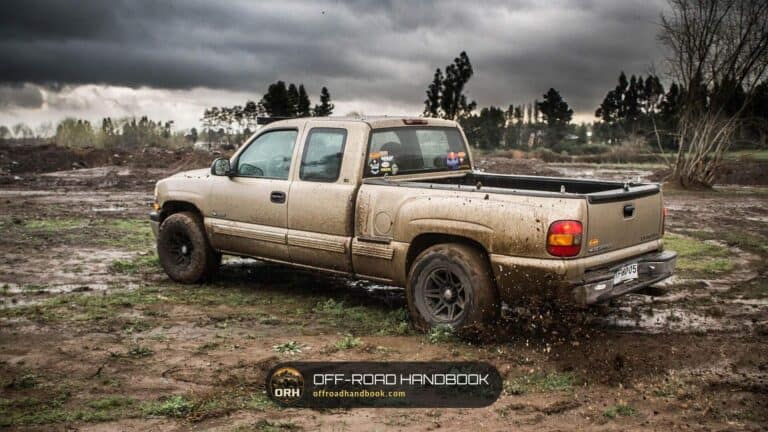 Muddy pickup truck kicking up dirt on a wet, rutted off-road track under a stormy sky