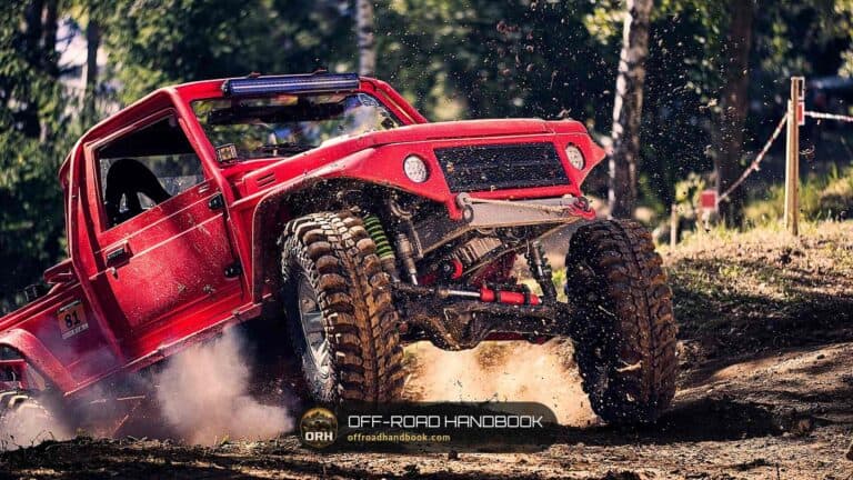 Red off-road 4x4 with big mud tires kicking up dirt on a trail, one front wheel lifted