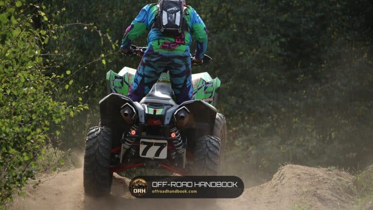 ATV rider standing on the footpegs of a green quad, seen from behind, on a dirt track through dense green woods