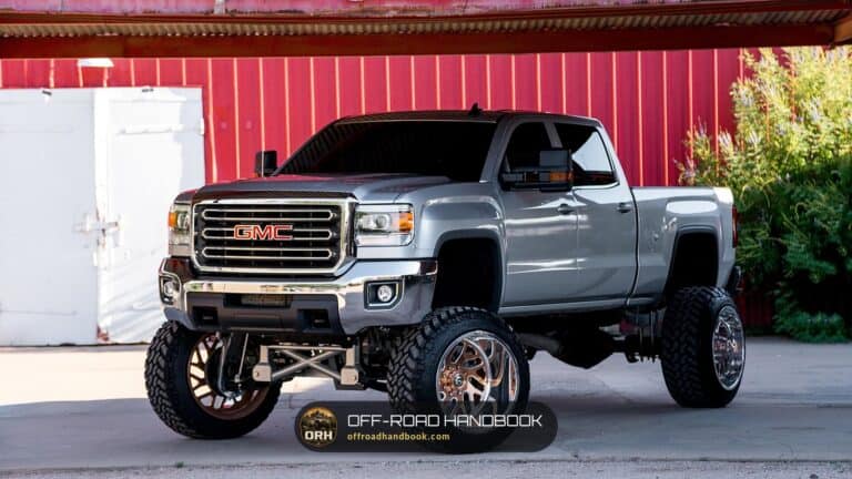 Silver lifted heavy-duty pickup with large wheels and off-road tires parked in front of a red building