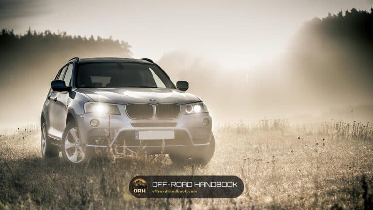 Silver crossover SUV with headlights on driving on a dusty dirt track through dry grass in morning haze