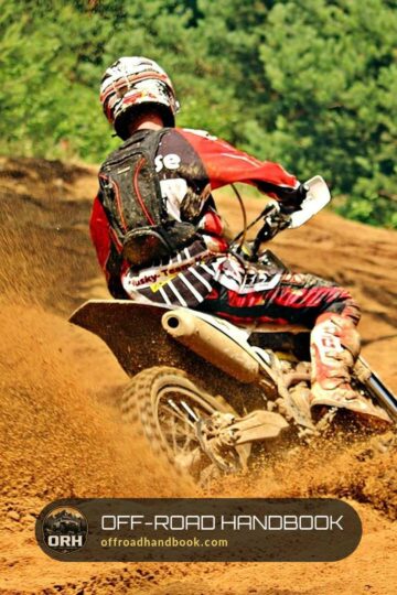 Dirt bike rider in a red jersey leaning through a sandy berm on a wooded trail, throwing sand off the rear tire