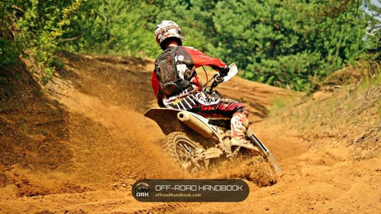Dirt bike rider in a red jersey leaning through a sandy berm on a wooded trail, throwing sand off the rear tire
