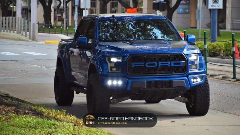 Blue Ford F-150 pickup with aftermarket fog lights and large off-road tires at a curb