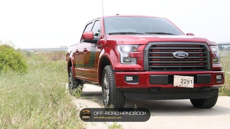 Red Ford F-150 crew-cab pickup parked on a dirt path beside tall grass