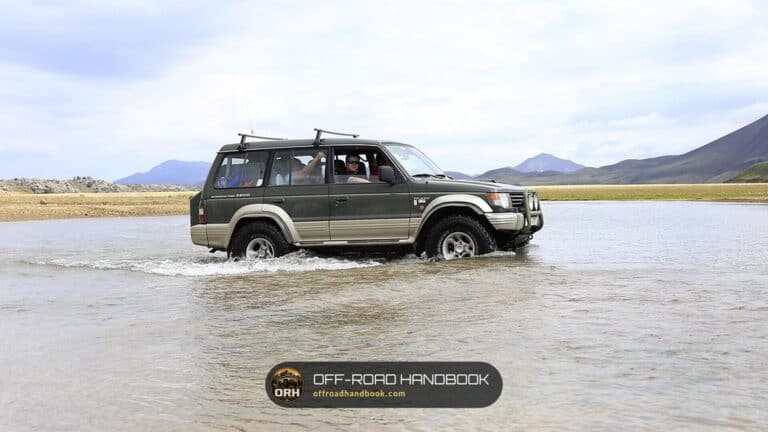 Green and tan SUV driving through shallow braided river water with mountains behind it