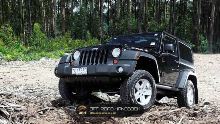 Black Jeep Wrangler Rubicon parked on a dusty forest clearing with tall trees behind it