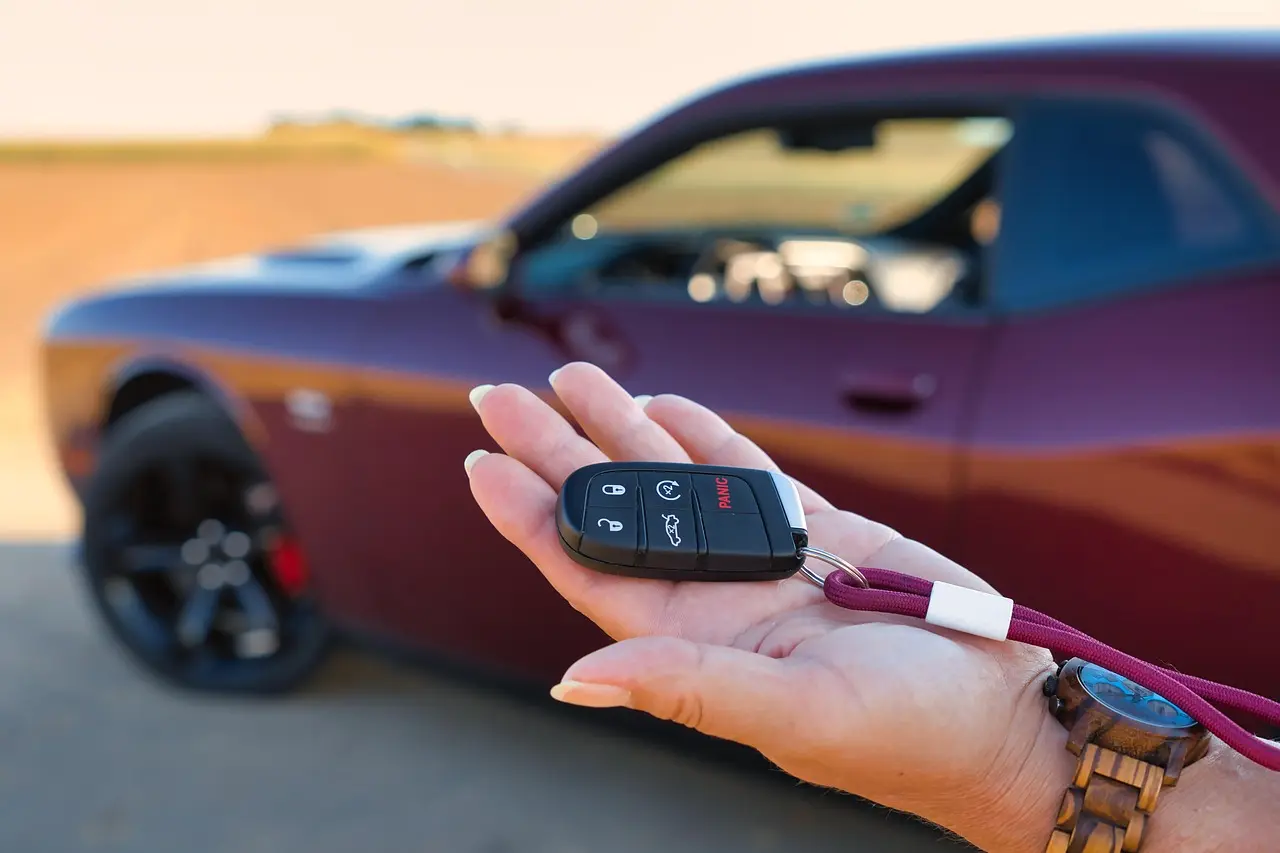Hand holding a Chrysler-family key fob with lock, unlock and panic buttons in front of a parked car