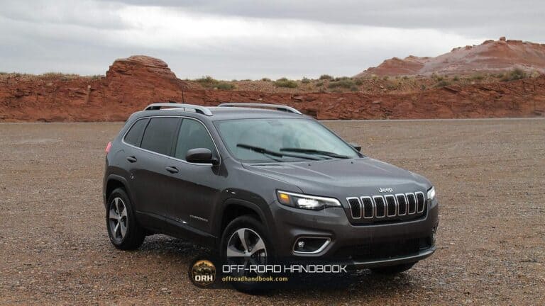Gray Jeep SUV with the seven-slot grille parked on a gravel lot with red rock hills behind