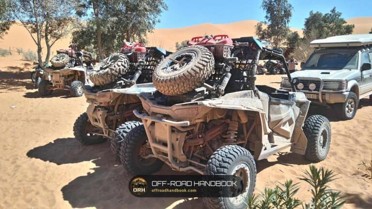 Rear view of three dusty side-by-side off-road vehicles with spare tires and fuel cans parked on desert sand