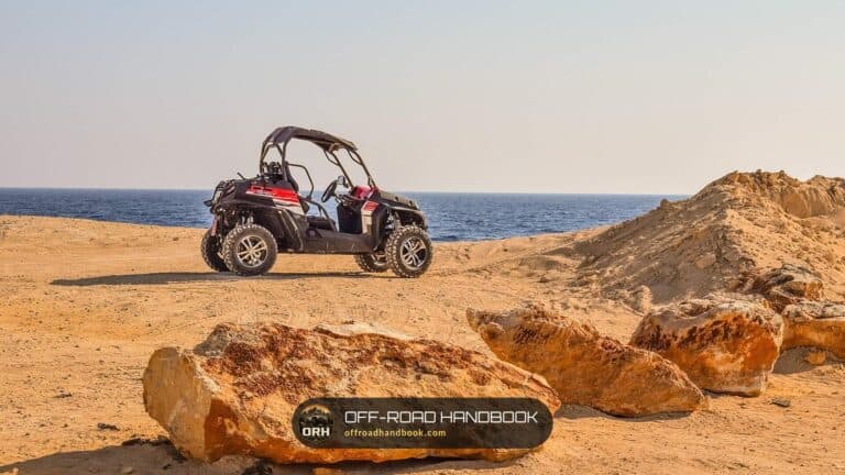 Red and black side-by-side utility vehicle parked on a sandy dirt area beside the sea