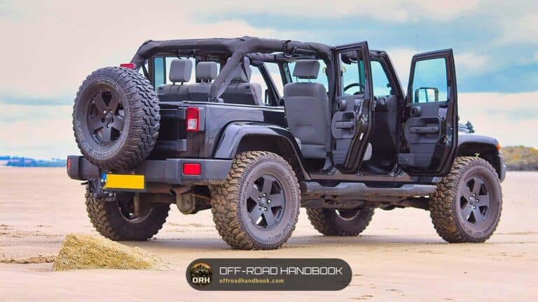 Black four-door Jeep Wrangler Unlimited with its doors removed, parked on a sandy beach on muddy off-road tires