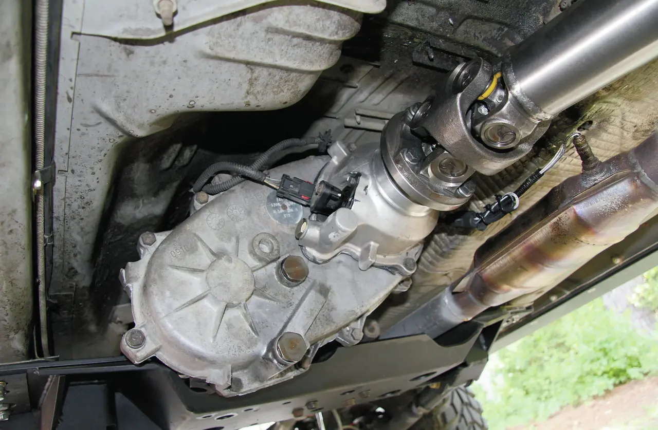 Power transfer unit bolted to the transmission on a Jeep Cherokee, seen from underneath with the driveshaft and exhaust beside it