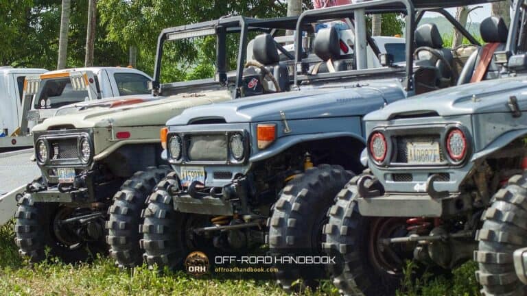 Three lifted vintage four-wheel-drive trucks with large off-road tires parked in a row on grass