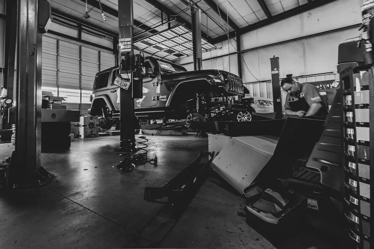 Black and white shop photo of a Jeep Wrangler raised on a lift with its suspension hanging while a mechanic works nearby