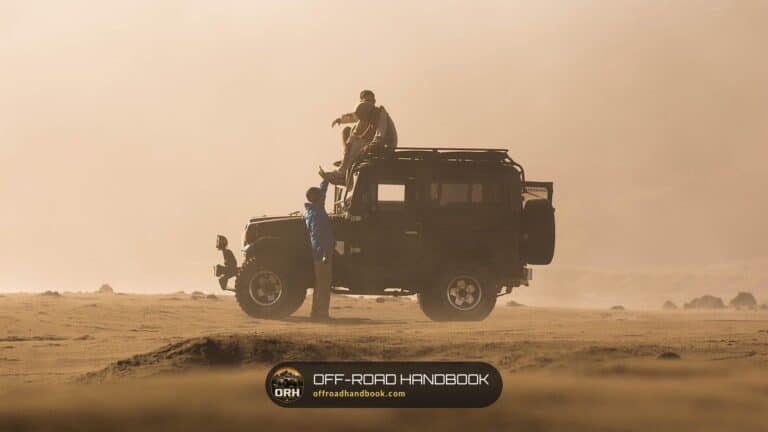 A dusty four-wheel-drive truck with passengers parked on a hot desert track in a sandstorm haze