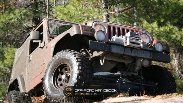 Muddy lifted Jeep Wrangler-style 4x4 with big mud tires and a winch bumper on a rocky forest trail, seen from low in front
