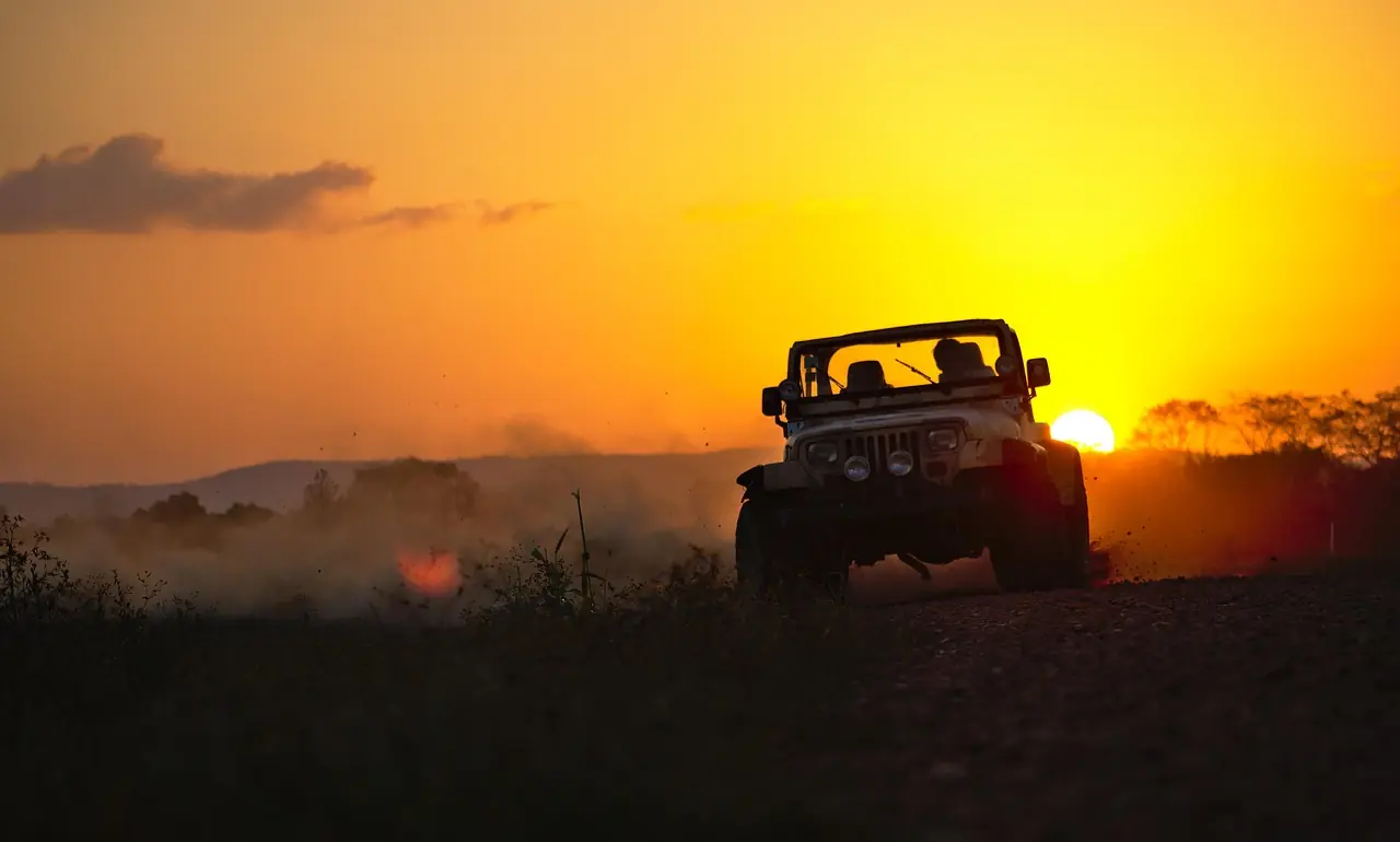 Open-top Jeep silhouetted on a dusty dirt road at sunset